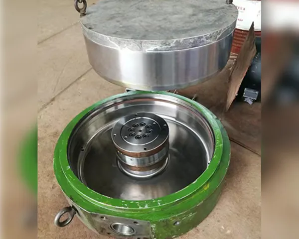 Round metal cylindrical mechanical component with green housing and silver internal parts resting on a pale concrete floor, soft daylight tones.