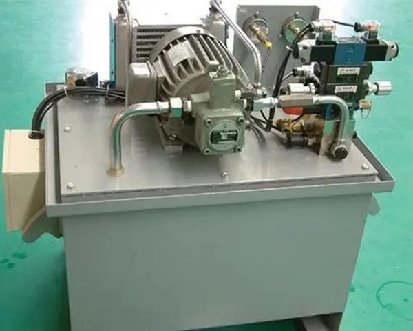 Small industrial hydraulic power unit resting on a green workshop floor, with a soft teal background, neutral gray components, and clean overhead light.